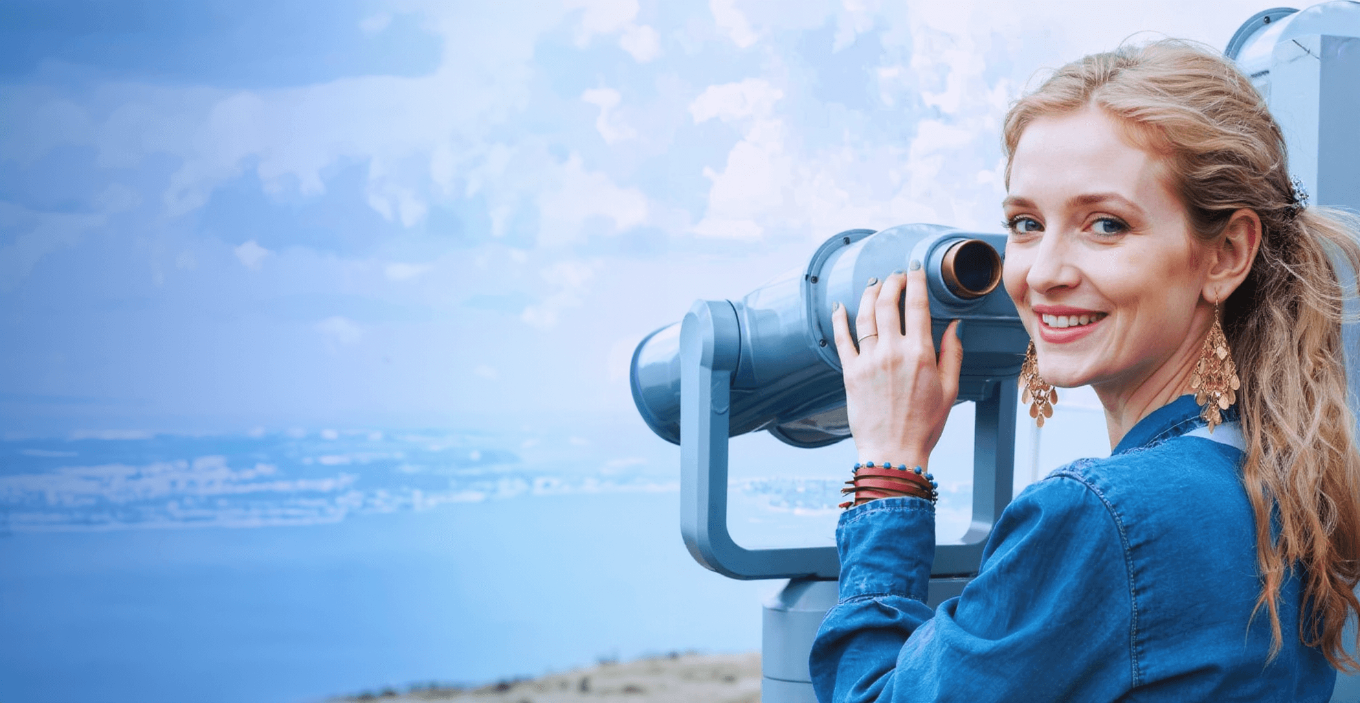 A women standing on the shore next to a large telescope and looking back towards the camera